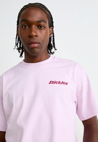 Šviesiai rožinė medvilninė marškinėliai, kuriuose yra raudonas grafikos logotipas su užrašu "Dickies" kairėje krūtinės pusėje. Trumpomis rankovėmis ir apvaliu kaklu.