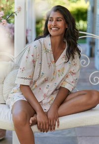 Floral-printed, light beige cotton loungewear set with short sleeves, button closure, and lace accents, displayed on a seated model.
