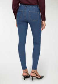 Kobieta w dopasowanych niebieskich dżinsach, bordowej bluzce z długim rękawem i czarnych szpilkach typu slingback, ukazana od tyłu na białym tle.