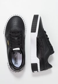 Black leather sneakers with perforated texture, black laces, white rubber sole, and gold logo accent on the side. Flat design with rounded toe.
