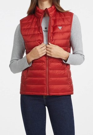 Kobieta w czerwonej pikowanej kamizelce bez rękawów na jasnoszarej, długorękawowej koszulce, zapinająca przedni suwak, ubrana w ciemnoniebieskie dżinsy.