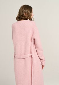 Langer pinker Cardigan aus weichem, strukturiertem Stoff. Besitzt einen Gürtel in der Taille und hat einen lockeren Schnitt mit einem mittellangen Design.