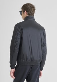 Giacca a sacco nera con colletto alto, polsini a costine e orlo elastico. Texture liscia e design leggero, vista da dietro.