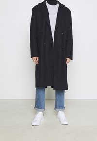 Cappotto nero doppiopetto con colletto a scollo, indossato sopra un dolcevita grigio, abbinato a jeans azzurri chiari con risvolto e sneakers bianche.