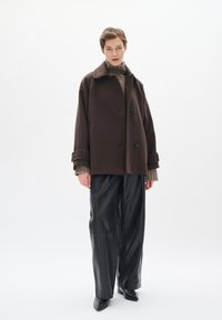 Abrigo de lana marrón con cuello, que presenta dos botones frontales y puños grandes. Combinado con pantalones de cuero negros y un suéter texturizado.