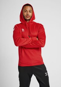 Rote Kapuzenjacke aus glattem Stoff, mit einem Logo auf der Brust und kontrastierenden weißen Akzenten an den Ärmeln.