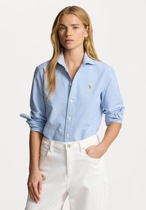 Light blue button-up shirt with long sleeves, rolled at the cuffs, featuring a small embroidered logo on the chest. Worn with white pants.