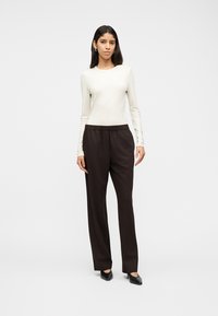 Γυναίκα που στέκεται φορώντας εφαρμοστό κρεμ ριμπ μπλούζα με μακριά μανίκια, φαρδιά μαύρη παντελόνια και μαύρα παπούτσια με τακούνι, μπροστά σε απλό φόντο.