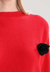Rode geribbelde sweater met een zwarte vilten roosaccent aan de linkerkant van de borst. Zachte textuur en regulier pasvorm ontwerp.