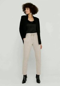 Bež hlače z vrvico, narejene iz mehkega blaga, v kombinaciji s črnim blejzerjem in črno satensko zgornjo obleko. Model nosi črne gležnjarje.