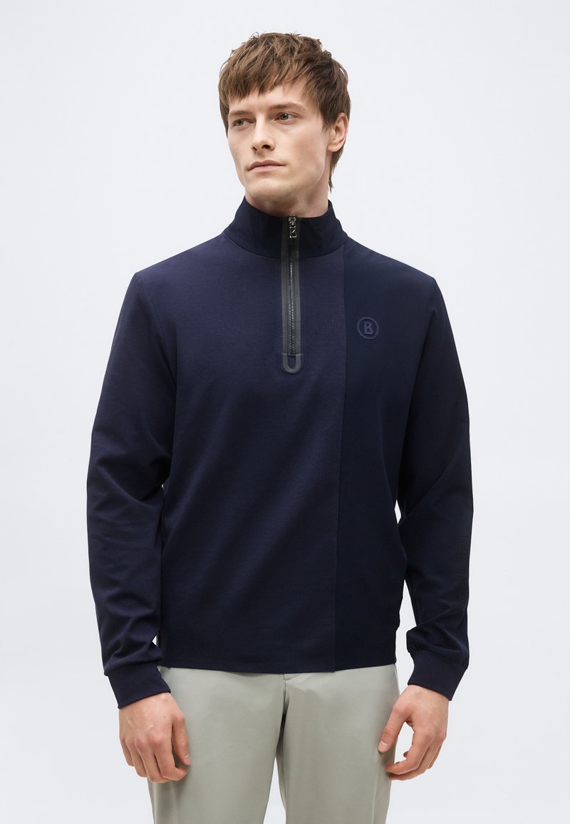 Uomo che indossa un maglione blu navy con zip e collo alto, e pantaloni grigio chiaro, in piedi davanti a uno sfondo bianco semplice.