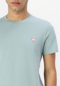 Lyseblå bomulds T-shirt med korte ærmer, der har et lille rødt trekantslogo på venstre bryst. Glat overflade og rund halsudskæring.