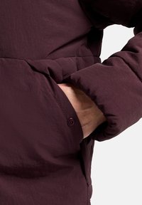 Bordeauxkleurige pufferjack met een textuurstof. Heeft een zijzak met een knoopsluiting. Zichtbare hand gedeeltelijk in de zak gestoken.