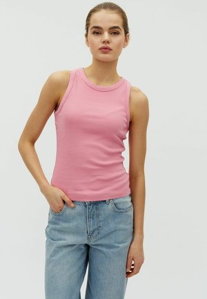 Ung kvinde iført en lyserød ærmeløs top og lyseblå jeans, stående med den ene hånd i lommen foran en ensfarvet baggrund.