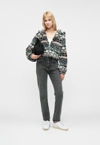 Mikina s kapucňou z fleecu s mnohofarebným geometrickým vzorom, čierne džínsy a biele tenisky. Model drží čiernu tašku.