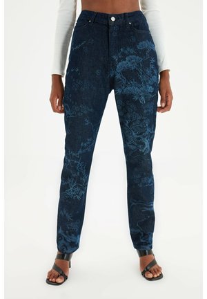 Donkerblauwe high-waist jeans met lichtblauwe bloemmotieven, gedragen met zwarte sandalen met hak en een wit cropped top met lange mouwen.