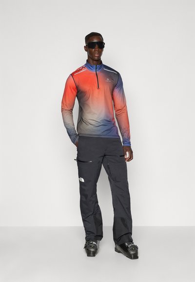 Sudadera de manga larga con cremallera en un degradado de rojo a azul. Combinada con pantalones impermeables negros con bolsillos con cremallera y logo. Calzado negro.