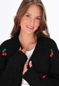 Juodas megztinis su raštų struktūra, dekoruotas raudonais vyšnių siuvinėjimais. Modelis dėvi šviesią viršutinę dalį po juo.