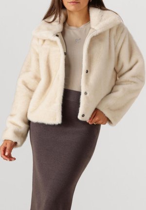 Crème neplederen jasje met een kraag, vooraan drukknopen en lange mouwen, gedragen over een geribbelde beige top en een donkergrijze aansluitende rok.
