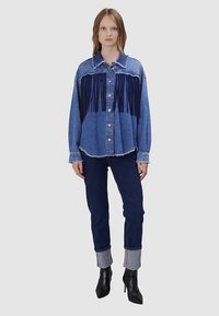 Chemise en denim bleu avec détail à franges sur la poitrine, associée à un pantalon sombre avec revers retroussés et des bottines noires à talons.