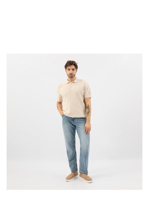 Man met tatoeages, gekleed in een beige poloshirt, blauwe spijkerbroek en beige instapschoenen, staat met één hand in de zak tegen een witte achtergrond.