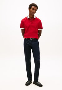 Rdeča polo majica z kontrastnimi belimi in mornarsko modrimi poudarki na ovratniku, kombinirana z mornarsko modrimi hlačami ozek kroj in črnimi čevlji. Minimalističen dizajn.