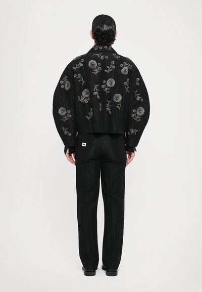 Schwarze bestickte Jacke mit floralen Mustern, entspannte Silhouette und ausgestellt geschnittene Ärmel. Kombiniert mit passenden schwarzen Hosen und Schuhen.