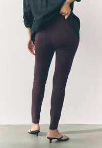 Bordeaux skinny jeans met een soepele textuur, voorzien van twee achterzakken en een middelhoge taille. Gecombineerd met zwarte slipper-sandalen.