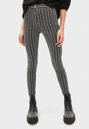 Femme portant un pantalon rayé vertical noir et blanc, une ceinture noire et des bottines à plateforme noires cloutées sur fond uni.