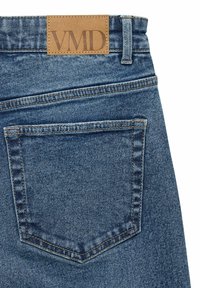 Modra denim hlače z rjavim usnjenim znakom "VMD" na pasu. Imajo en zadnji žep z vidnimi šivi.