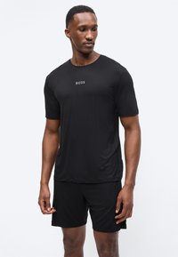 Άνδρας με μαύρο αθλητικό σορτς και μαύρο μπλουζάκι BOSS, στέκεται με χαλαρά τα χέρια, κοιτώντας προς τα κάτω και αριστερά, μπροστά σε λευκό φόντο.