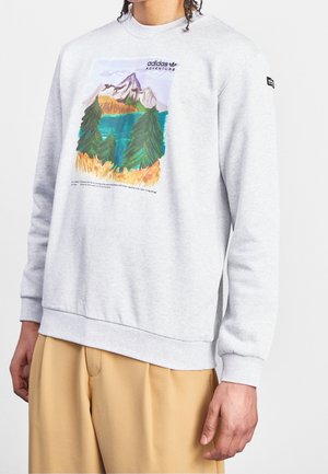 Person iført en lysegrå sweatshirt med farverig grafik af bjerge og skov samt teksten "adidas Adventure" foran, kombineret med beige bukser.