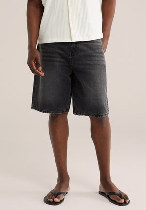 Muž oblečený v čiernych rifľových šortkách, bielej krátkorukávovej košeli na gombíky a čiernych žabkách, stojaci pred jednoduchým pozadím.