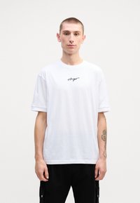 Hvid bomulds T-shirt med korte ærmer, prydet med et sort håndskrevet logo på brystet. Glat tekstur og afslappet pasform.