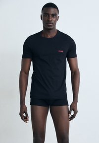 T-shirt noir à manches courtes en coton, arborant un petit logo rouge "HUGO" sur la poitrine gauche, porté avec un boxer noir.
