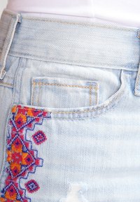 Vaaleansiniset farkut, joissa on moniväristä kirjontaa etutaskun ympärillä ja kulutustahelman lähellä.