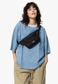 Marsupio nero in tessuto indossato a tracolla su una camicia di jeans oversized azzurra con maniche arrotolate e pantaloni beige casual.