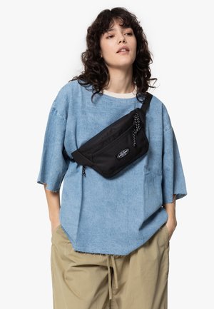Un sac banane noir porté en travers du corps sur une chemise en denim oversize bleu clair, avec des manches retroussées, et un pantalon beige décontracté.