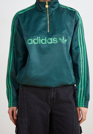Veste de survêtement Adidas vert foncé avec des bandes vert clair sur les manches, fermeture éclair dorée à moitié, et logo vert clair sur la poitrine, portée avec un pantalon noir.