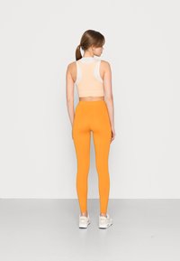 Reggiseno sportivo beige con accenti bianchi, abbinato a leggings arancioni brillanti. Texture liscia, design aderente e sneakers bianche alte fino alla caviglia.