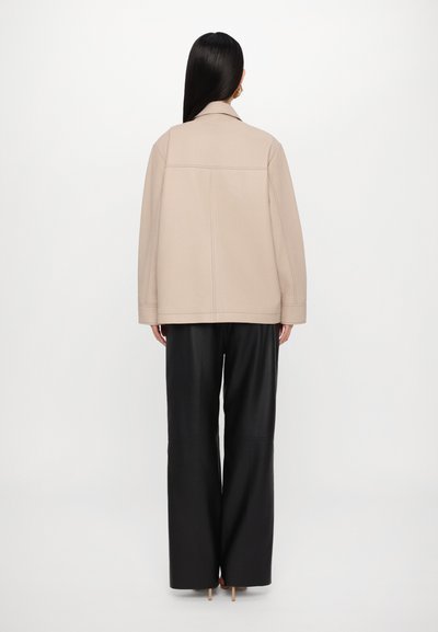 Jachetă oversized bej cu guler, detalii cusute și buzunare laterale, asociată cu pantaloni negri cu talie înaltă și picioare largi.