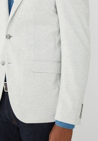 Hellgrauer Blazer aus strukturiertem Stoff, mit einer Brusttasche, zwei Knöpfen und Bündchen mit drei Knöpfen. Darunter ein blaues Hemd.