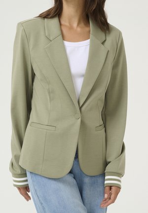Femme portant un blazer vert clair ajusté avec des poignets rayés sur un haut blanc et un jean bleu clair, posant devant un fond uni.