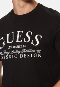 Camiseta de algodón negra con gran texto impreso en blanco, que incluye "GUESS", "LOS ÁNGELES, CA" y "DISEÑO CLÁSICO". Cuello redondo, mangas cortas.