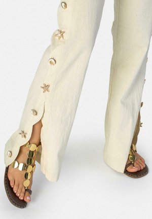 Pantalones beige de pierna ancha con botones de concha marina y estrella de mar a lo largo de los lados, combinados con sandalias doradas de disco circular sobre pies descalzos.