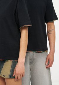 Δύο άτομα που φορούν μαύρα T-shirts με καμουφλάζ περιθώριο, το ένα με φούστα πατσγουόρκ από τζιν και το άλλο με ανοιχτό γκρι τζιν.