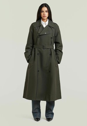 Junge Frau trägt einen langen, dunkelgrünen Trenchcoat mit Gürtel über einem weißen Hemd und blauen Jeans, steht mit den Händen in den Manteltaschen.