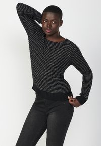 Maglione nero e argento con texture, scollatura ampia, maniche lunghe e orlo a coste, abbinato a jeans neri. Sfondo neutro.