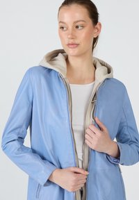 Vaaleansininen nahkatakki, jossa on vaaleanbeige huppari vuoritus, vetoketjukiinnitys edessä, sivutaskut ja pitkät hihat. Sujuva pinta ja moderni muotoilu.