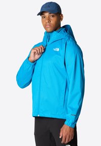 Svetle modrá nepromokavá bunda s předním zipem, nastavitelnou kapucí a elastickými manžetami, na hrudi má malé bílé logo.